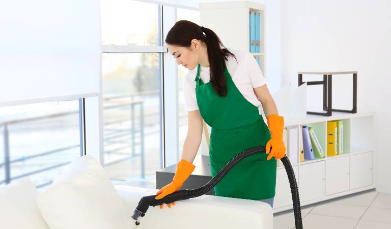 Young woman in white-green dress vacuuming sofa inside a office