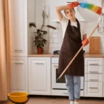 Woman in uniform holding a duster and cleaning stick in her hand inside her kitchen.