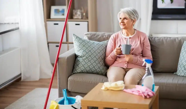 Old lady on sofa drinking coffee with cleaning tools around her.