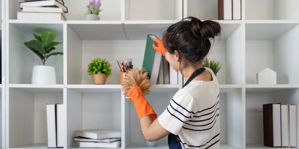 A lady in orange gloves cleaning books cabinet.