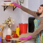 Young man in the kitchen wearing apron and pink gloves cleans the shelf