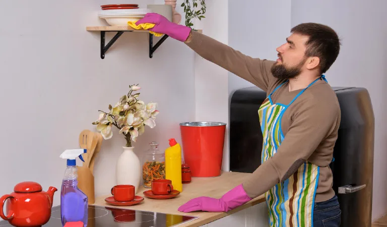 Young man in the kitchen wearing apron and pink gloves cleans the shelf