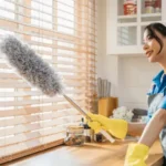 Asian woman with duster cleans dirty window blinds enjoying housework ensuring hygiene for a modern clean home