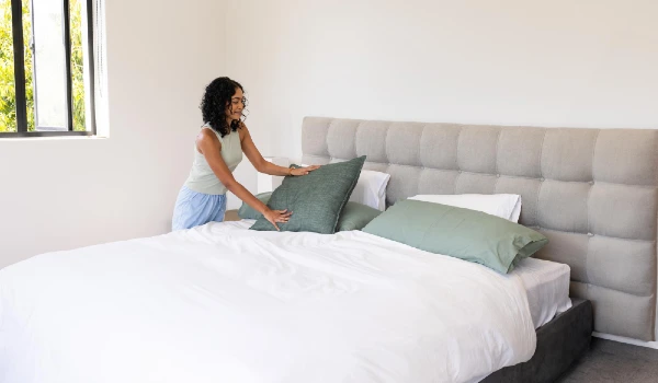 Woman lifting green pillow onto grey headboard in bedroom by black framed window
