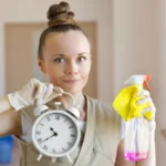 Young cleaning lady holding cleaning spray and a clock in the corridor
