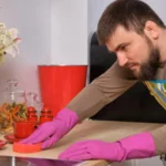 Bearded man in the kitchen, wearing apron and pink gloves cleans the cooking surface using detergents