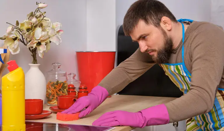 Bearded man in the kitchen, wearing apron and pink gloves cleans the cooking surface using detergents